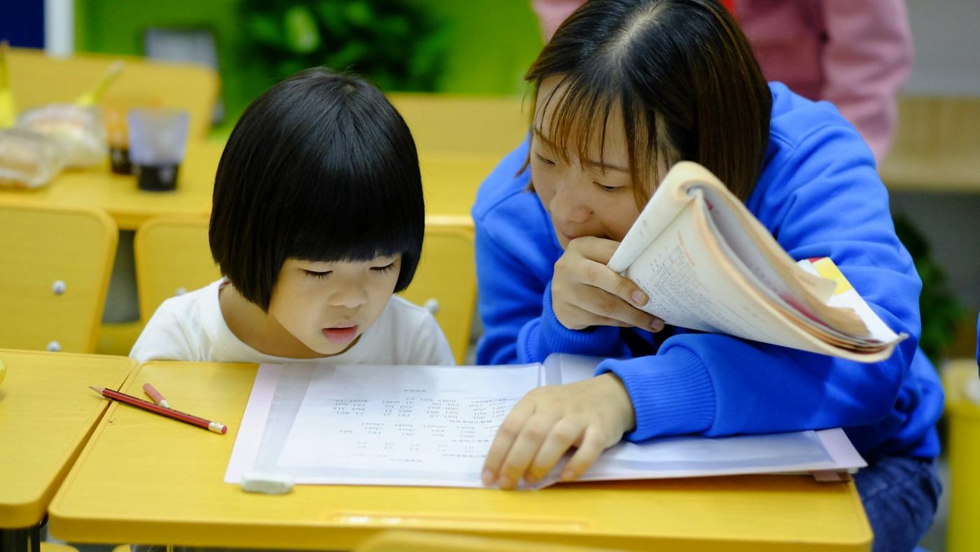 A guide leans in to help a young girl with her work at a yellow table