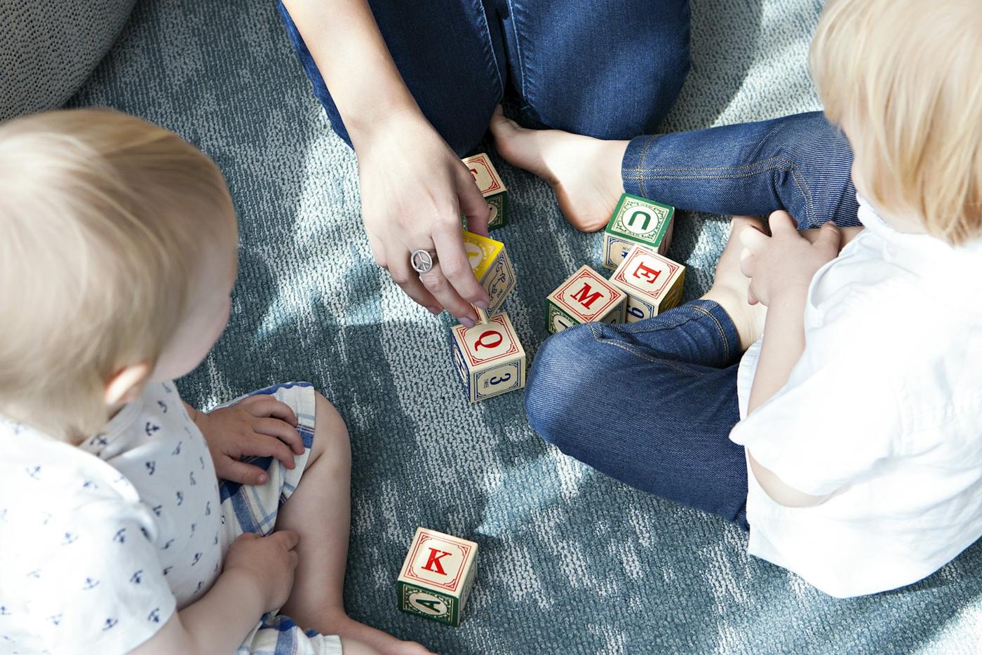 Two toddlers play with wooden letter cubes on a rug